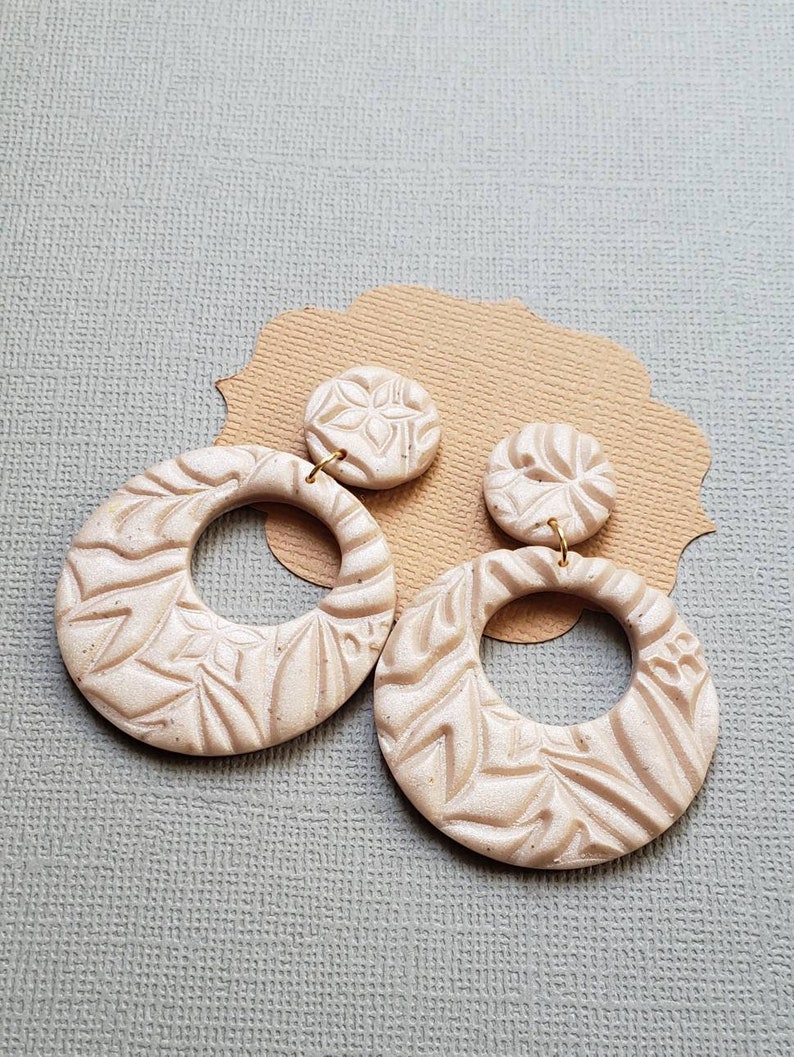 May include: A pair of beige, textured earrings. Each earring features a circular design with a floral pattern. The earrings are attached to small, round studs with a similar design. The earrings are displayed on a brown, decorative paper cutout.