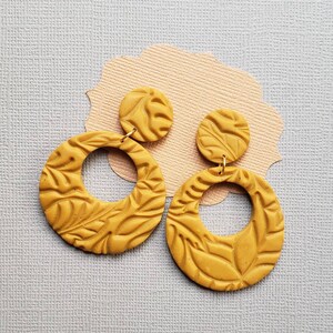 May include: Mustard yellow earrings with a leaf pattern. The earrings have a circular design with a smaller, round stud at the top. The earrings are made of a clay-like material and are displayed on a light gray surface.