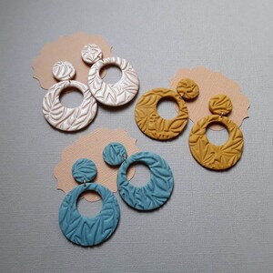 May include: Four pairs of handmade earrings in various colors. The earrings feature a circular design with a textured leaf pattern. Colors include white, mustard yellow, and teal. Each earring has a small stud and a larger hoop, all made of clay.