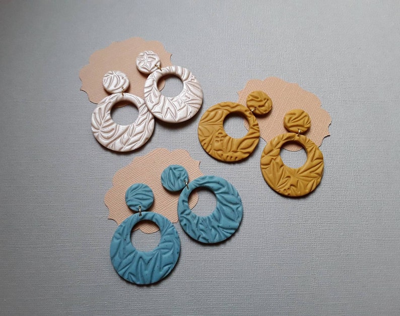 May include: Four pairs of handmade earrings in various colors. The earrings feature a circular design with a textured leaf pattern. Colors include white, mustard yellow, and teal. Each earring has a small stud and a larger hoop, all made of clay.