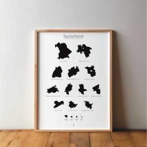 May include: A framed print featuring a minimalist map of Germany, showcasing the outlines of its states in black against a white background. The word "Deutschland" is at the top. The print is displayed on a wooden surface.