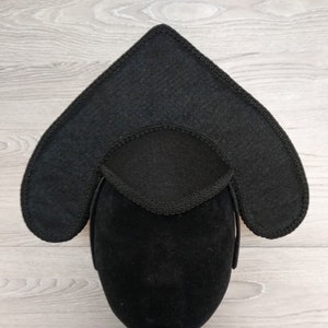 May include: A black, heart-shaped hat with a decorative trim. The hat is made of a dark, textured fabric and is displayed on a black mannequin head. The hat's design includes a central, rounded section and two extended, pointed sides.
