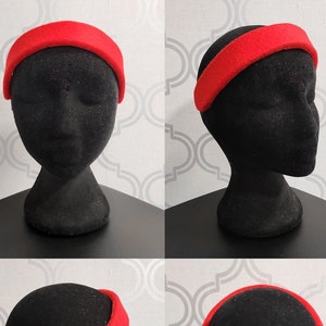 May include: A bright red headband with a wide, padded design. The headband is shown on a black mannequin head, with a black elastic band at the back. The headband is a solid color and appears to be made of felt or a similar fabric.