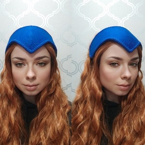 May include: A vibrant blue felt headband with a distinctive V-shaped front. The headband is a solid color and appears to be handmade. The image shows three views of the headband, showcasing its design and shape. This accessory is likely for fashion or costume purposes.