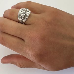May include: A wide silver ring crafted from a repurposed spoon, featuring an intricate embossed floral and scrollwork pattern across the entire outer surface. The metal has a polished finish with darkened recessed areas that highlight the detailed design.