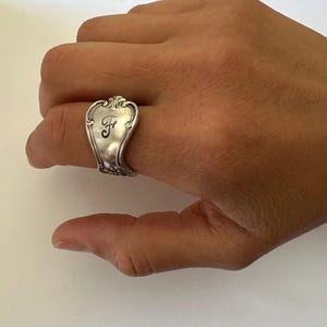 May include: A silver-toned ring crafted from a repurposed spoon handle, featuring an ornate, curved shape with decorative embossed detailing along the edges and a prominent, elegant script letter F engraved in the center of the metal surface.