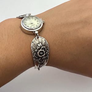May include: A silver-toned quartz wristwatch featuring a round white face with black numerals and hands. The watch band is crafted from a repurposed silver-colored metal spoon handle, showcasing an embossed floral pattern with detailed petals.
