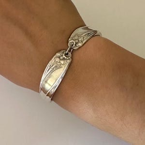 May include: Silver spoon bracelet with floral detailing. The bracelet is made from repurposed silverware, featuring a curved design with a flower motif. The bracelet is connected with a small chain and is worn on a wrist.