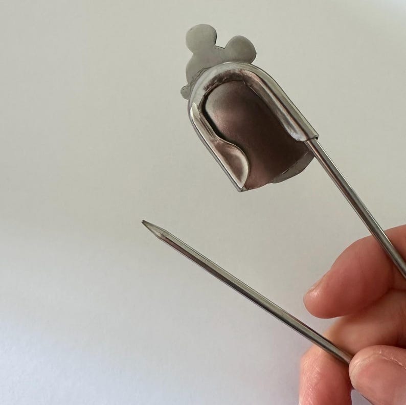 Pu&ograve; includere: Un tappo per bottiglia in metallo color argento con un design a forma di Mickey Mouse sulla parte superiore. Il tappo ha un corpo a forma di campana e un lungo perno appuntito. L'immagine &egrave; un primo piano.
