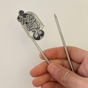 May include: A silver-toned metal kilt pin featuring a raised, embossed character design of a cartoon mouse wearing a bow and dress. The pin has a long, pointed needle-like shaft and a secure clasp mechanism at the top, crafted from polished metal.