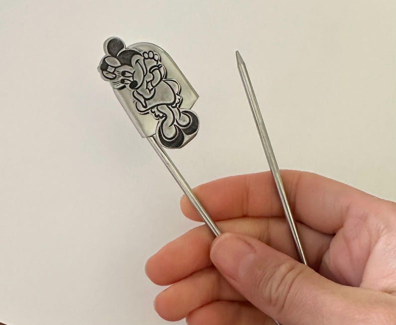 May include: A silver-toned metal kilt pin featuring a raised, embossed character design of a cartoon mouse wearing a bow and dress. The pin has a long, pointed needle-like shaft and a secure clasp mechanism at the top, crafted from polished metal.