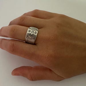 May include: A wide silver-toned metal ring featuring an embossed owl design. The owl has detailed circular eyes, a pointed beak, and textured wing patterns covering the band. The metal has a darkened, oxidized finish in the recessed areas.