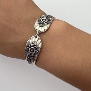 May include: A silver-toned bracelet constructed from two repurposed spoon handles. Each link features an embossed floral pattern with intricate petals and leaves, connected by a small metal jump ring to form a continuous circular band.