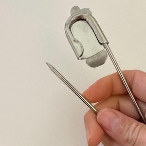 May include: A large silver-toned metal safety pin featuring a flat, shield-shaped decorative plate attached to the top coil. The pin is constructed from a single piece of wire with a sharp, pointed end and a secure clasp mechanism for fastening.