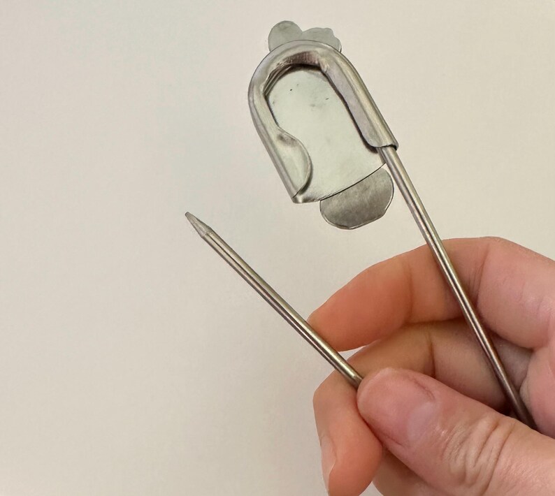 May include: A large silver-toned metal safety pin featuring a flat, shield-shaped decorative plate attached to the top coil. The pin is constructed from a single piece of wire with a sharp, pointed end and a secure clasp mechanism for fastening.
