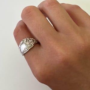 May include: A silver spoon ring with a floral design. The ring is worn on a finger, showcasing the intricate details of the repurposed silverware. The ring's design features a detailed floral pattern, adding a touch of vintage charm.