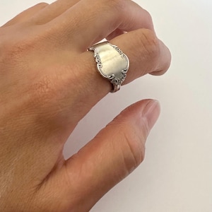 May include: A silver-toned ring crafted from a repurposed spoon handle, featuring an ornate, embossed floral pattern along the edges and a smooth, polished central surface that wraps around the finger in a curved, metallic band design.