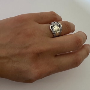 May include: A silver-toned ring crafted from the handle of a vintage spoon, featuring an ornate, embossed floral pattern with darkened recessed details. The metal band wraps around the finger, showcasing a polished finish and intricate scrollwork.