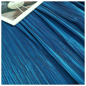 Puede incluir: Primer plano de una tela plisada en un vibrante azul turquesa. La tela tiene un aspecto texturizado y acanalado con líneas verticales. La imagen también muestra un objeto con marco blanco con hilo y agujas de tejer.