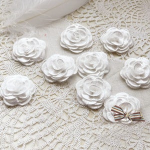 Puede incluir: Diez adornos de flores de tela blanca con textura rizada, dispuestos sobre una superficie de crochet blanca.