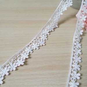 Puede incluir: Borde de encaje blanco con un delicado patrón floral. El borde presenta pequeñas flores espaciadas uniformemente con un borde festoneado.