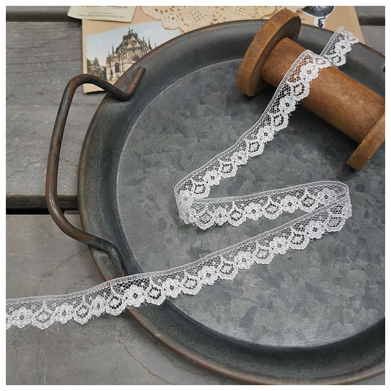 K&ouml;nnte beinhalten: Wei&szlig;e Spitzenborte mit zartem Blumenmuster. Die Borte ist auf einer Holzspule aufgerollt und auf einem Metalltablett ausgestellt.