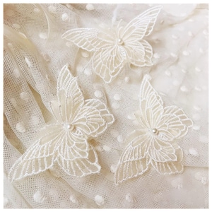 May include: Three ivory-colored, embroidered butterfly appliques with pearl centers are arranged on a white, dotted fabric. The butterflies have detailed wing patterns and are made of delicate, sheer material.