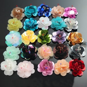 Może przedstawiać: Zestaw 21 kolorowych, kwiatowych ozdób z tkaniny z cekinami i perłowymi akcentami. Ozdoby są ułożone w rzędach na czarnej powierzchni.