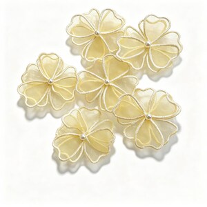 Könnte beinhalten: Eine Ansammlung von sechs blassgelben Organza-Blumenapplikationen, jede mit fünf durchscheinenden Blütenblättern mit genähten Rändern und einer kleinen weißen Perle in der Mitte jedes einzelnen Blumenelements, die in einer Gruppe angeordnet sind.
