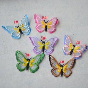 Puede incluir: Seis apliques de mariposa bordados en varios colores, incluyendo azul, rosa, morado, verde y marrón. Cada mariposa tiene un centro amarillo y está etiquetada con un número del 1 al 6.
