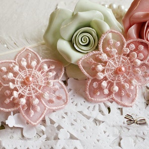 Può includere: Due decorazioni floreali in pizzo rosa con centri di perle, una piuma bianca e due rose artificiali, una verde e una rosa, su uno sfondo di pizzo bianco.