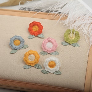 Könnte beinhalten: Sechs gestickte Blumenapplikationen in verschiedenen Farben, darunter Orange, Blau, Rosa, Gelb, Weiß und Grün. Jede Blume hat einen Mittelkreis und ist von Blütenblättern umgeben. Die Blumen befinden sich auf einem beigefarbenen Stoffhintergrund.