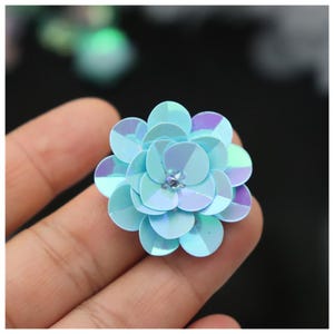 May include: A layered flower-shaped embellishment crafted from iridescent light blue sequins. The circular sequins are arranged in overlapping tiers to create a dimensional floral form, featuring a small clear bead at the center of the design.
