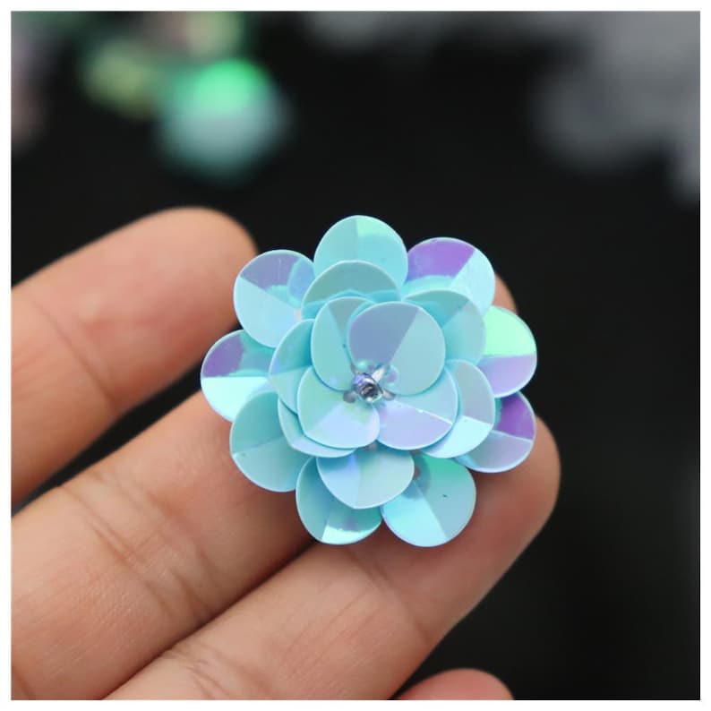 May include: A layered flower-shaped embellishment crafted from iridescent light blue sequins. The circular sequins are arranged in overlapping tiers to create a dimensional floral form, featuring a small clear bead at the center of the design.