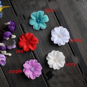 May include: Assortment of five fabric flower embellishments in various colors, including blue, white, ivory, red, and purple. Each flower has a pearl-like center.