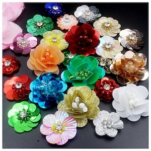 Op de afbeelding: Een verzameling kleurrijke, met pailletten versierde bloemen in verschillende maten en tinten, waaronder rood, groen, goud en blauw. Elke bloem heeft een centrale strass- of parelaccent, wat een sprankelend effect creëert. Ideaal voor knutselprojecten.