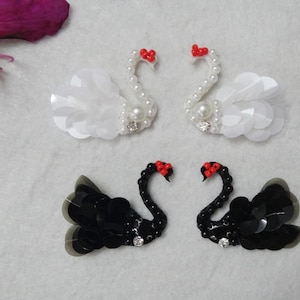 May include: Two pairs of swan-shaped embellishments, one pair white with pearl accents and one pair black with black sequins. Both pairs have red bows.