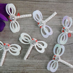 May include: Six white dragonfly embellishments with pearl accents and rhinestone centers. The dragonflies are arranged on a wooden surface.