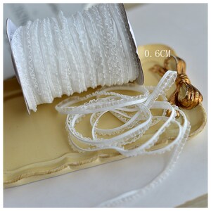 May include: White elastic lace trim with a ruffled edge. The trim is 0.6 inches wide and is shown on a white tray.