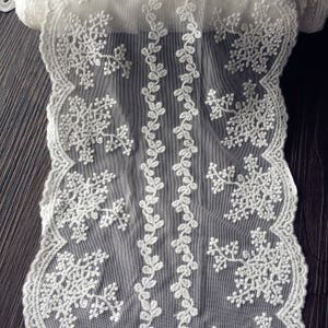 May include: Close-up of a white lace fabric with a delicate snowflake and floral design. The lace features a scalloped edge and a repeating pattern of snowflakes and vines. The fabric is likely used for decorative purposes.