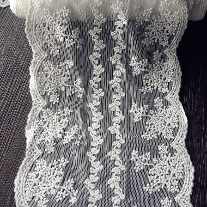 May include: Close-up of a white lace fabric with a delicate snowflake and floral design. The lace features a scalloped edge and a repeating pattern of snowflakes and vines. The fabric is likely used for decorative purposes.