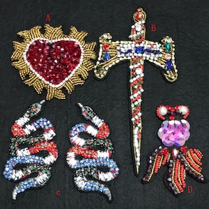May include: Four embroidered patches with jeweled embellishments. A red heart with gold rays, a jeweled sword, two snakes with red, white and blue scales, and a pink jeweled insect with red and gold accents.