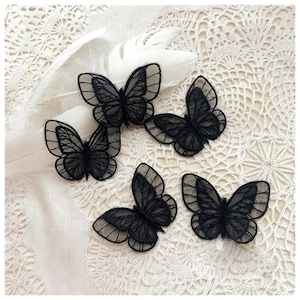 May include: Five black lace butterfly appliques with detailed wing patterns are arranged on a white lace surface, next to a white feather. The butterflies are intricately designed, showcasing a delicate, airy aesthetic.