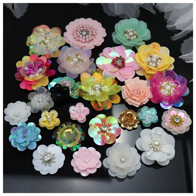 May include: A collection of assorted fabric flower appliques in various colors including pink, yellow, white, blue, and iridescent tones. Each flower features layered petals, some with sequin accents, central beads, crystals, or pearls.