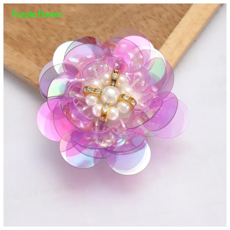May include: A decorative flower applique featuring layered translucent purple iridescent sequins arranged in a circular pattern. The center is embellished with a cluster of white faux pearls and gold-toned metal accents with clear crystals.