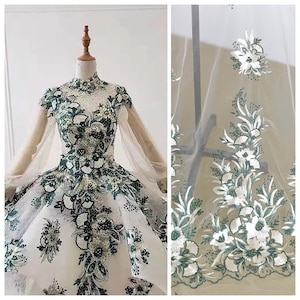 Puede incluir: Un vestido de novia de tul blanco con cuello alto y mangas largas. El vestido está decorado con bordados florales verdes y blancos.