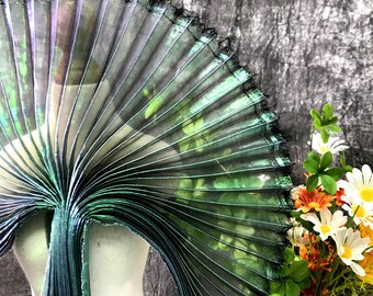 May include: A pleated, iridescent fabric in shades of green and black, with a hint of purple. The fabric is draped over a mannequin, with the text "black green magic color" visible. The edges are trimmed with black thread.