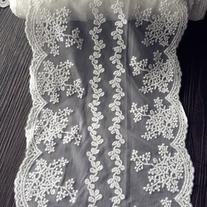 May include: Close-up of a white lace fabric with a delicate snowflake and floral design. The lace features intricate patterns and a scalloped edge. The fabric is likely used for crafting, sewing, or decorative purposes.