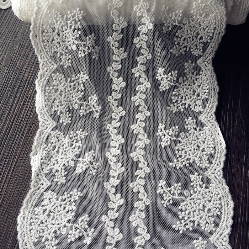 May include: Close-up of a white lace fabric with a delicate snowflake and floral design. The lace features intricate patterns and a scalloped edge. The fabric is likely used for crafting, sewing, or decorative purposes.