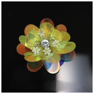 May include: A flower-shaped applique crafted from iridescent yellow and orange sequins. The center features a clear, faceted crystal surrounded by small, white, bead-like floral accents, creating a layered, reflective, and dimensional appearance.